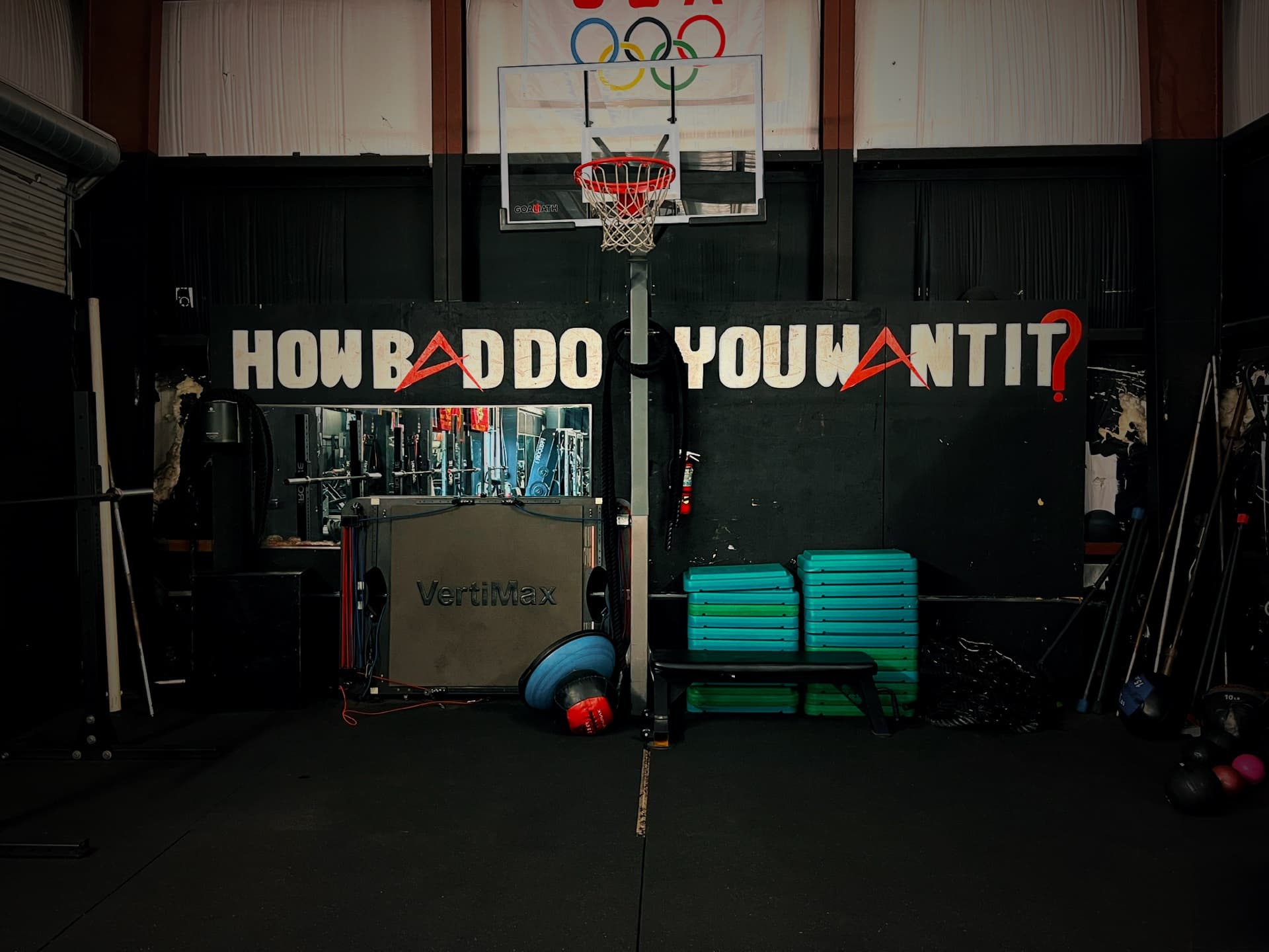 Black gym wall painted with the words How bad do you want it above a basketball hoop and medicine balls
